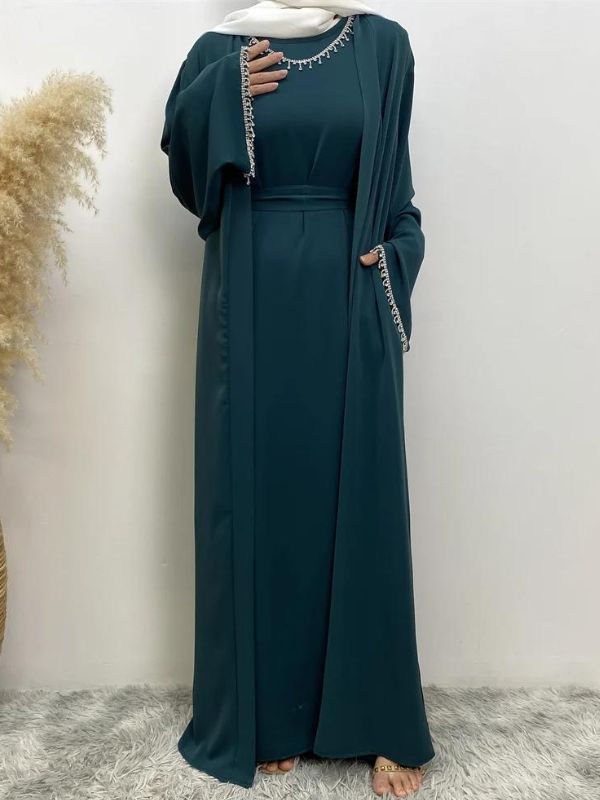 Femme portant une abaya vert émeraude avec des broderies argentées à l'encolure et aux manches, portant un hijab blanc, posant avec une main sur la poitrine et l'autre près de la taille, sur un fond neutre avec un vase de pampas à gauche