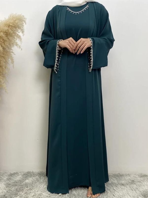 Femme portant une abaya vert émeraude avec des broderies argentées à l'encolure et aux manches, portant un hijab blanc, posant avec une main sur la poitrine et l'autre près de la taille, sur un fond neutre avec un vase de pampas à gauche