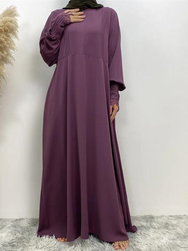 Une femme portant une abaya violette vue de face. La robe longue et fluide atteint le sol et présente des détails subtils tels que des plis sur le haut de la robe et des manches longues avec des motifs délicats. La femme porte également un hijab vert olive, qui complète harmonieusement la couleur de l'abaya. Le contraste entre le violet de la robe et le vert du hijab crée un ensemble visuellement attrayant