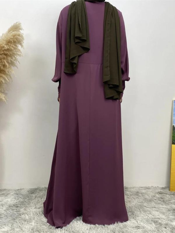 Une femme portant une abaya violette vue de face. La robe longue et fluide atteint le sol et présente des détails subtils tels que des plis sur le haut de la robe et des manches longues avec des motifs délicats. La femme porte également un hijab vert olive, qui complète harmonieusement la couleur de l'abaya. Le contraste entre le violet de la robe et le vert du hijab crée un ensemble visuellement attrayant