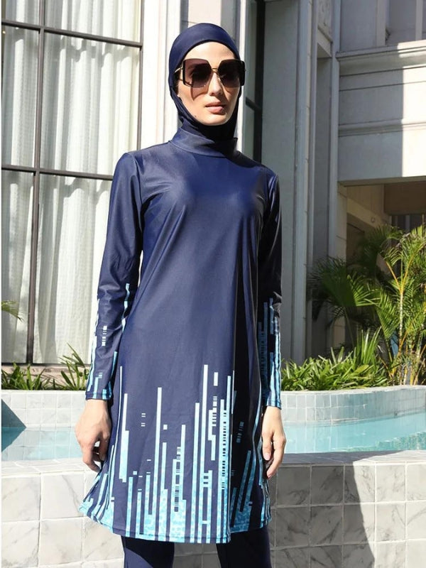 Une femme vêtue d'un burkini bleu marine avec des motifs géométriques bleu clair le long des manches et du bas de la tunique. Elle se tient près d'une piscine, portant des lunettes de soleil et regardant droit devant elle. Le design moderne du burkini, avec ses motifs verticaux stylisés, crée un contraste élégant avec le cadre environnant. Le décor de fond, composé d'un bâtiment classique et de végétation luxuriante, met en valeur l'ensemble tout en ajoutant une touche sophistiquée à la scène