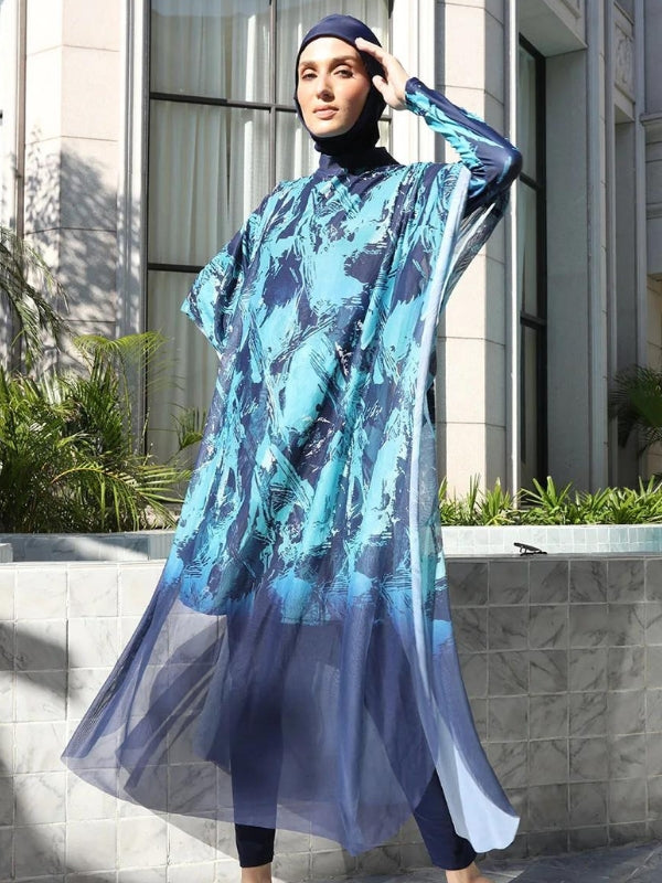 Une femme porte un burkini bleu avec des motifs abstraits dans des tons plus clairs de bleu. Le tissu fluide et léger de la tunique donne un effet éthéré, surtout au niveau de la jupe qui s'évase légèrement. Le voile bleu assorti complète la tenue, couvrant ses cheveux. Elle se tient dans un cadre moderne avec un mur carrelé et des plantes en arrière-plan, ce qui ajoute une touche contemporaine à l'ensemble