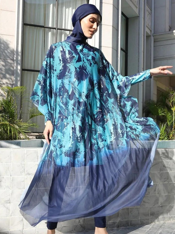 Une femme porte un burkini bleu avec des motifs abstraits dans des tons plus clairs de bleu. Le tissu fluide et léger de la tunique donne un effet éthéré, surtout au niveau de la jupe qui s'évase légèrement. Le voile bleu assorti complète la tenue, couvrant ses cheveux. Elle se tient dans un cadre moderne avec un mur carrelé et des plantes en arrière-plan, ce qui ajoute une touche contemporaine à l'ensemble