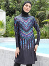 Une femme porte un burkini noir orné de motifs colorés dans des tons de bleu, violet et rose, formant un design géométrique en forme de V qui accentue la partie supérieure de la tenue. Le motif se prolonge vers le bas, créant un effet allongé et élégant. Elle est coiffée d&#39;un voile noir qui complète harmonieusement la tenue.