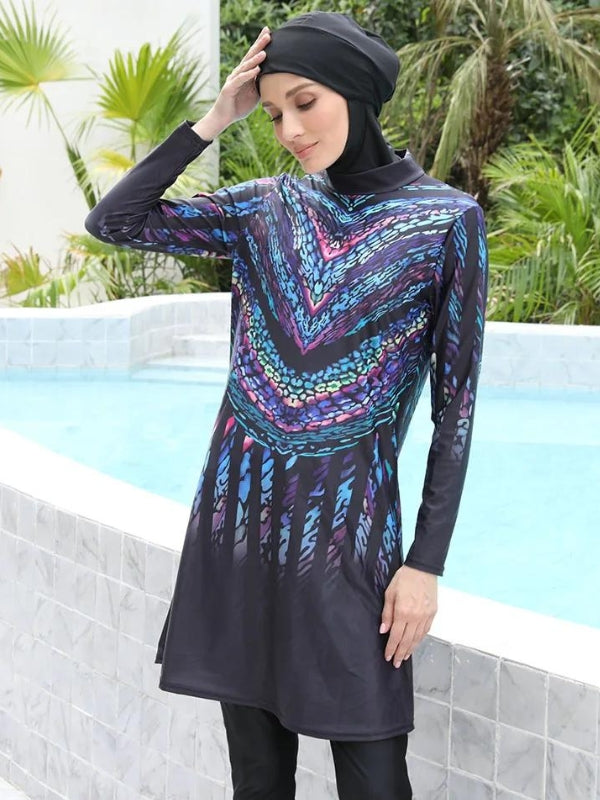 Une femme porte un burkini noir orné de motifs colorés dans des tons de bleu, violet et rose, formant un design géométrique en forme de V qui accentue la partie supérieure de la tenue. Le motif se prolonge vers le bas, créant un effet allongé et élégant. Elle est coiffée d'un voile noir qui complète harmonieusement la tenue.