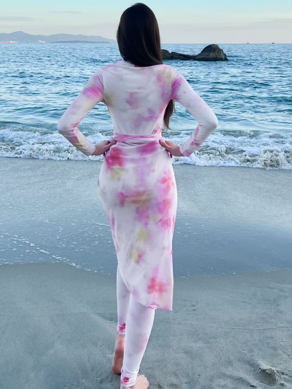 Une femme en burkini tie-dye rose et blanc, posant sur une plage avec les vagues à ses pieds