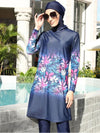 Une femme est photographiée portant un burkini bleu marine orné de motifs floraux vibrants dans des tons de rose, violet et vert. La tenue, composée d&#39;une tunique longue et de leggings assortis, offre une combinaison à la fois élégante et moderne. La femme complète son look avec un hijab assorti et de grandes lunettes de soleil. Elle pose à côté d&#39;une piscine, entourée d&#39;un décor extérieur luxueux avec de la verdure et des éléments architecturaux classiques en arrière-plan, créant une ambiance chic