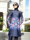 La femme sur cette photo porte un burkini bleu marine avec un motif floral coloré. La tunique est longue, descendant jusqu&#39;aux cuisses, et présente un dégradé de fleurs roses, violettes et vertes qui commence au milieu de la tenue et s&#39;étend vers le bas. Elle porte un hijab assorti, également bleu marine, ainsi que des lunettes de soleil qui ajoutent une touche de style supplémentaire