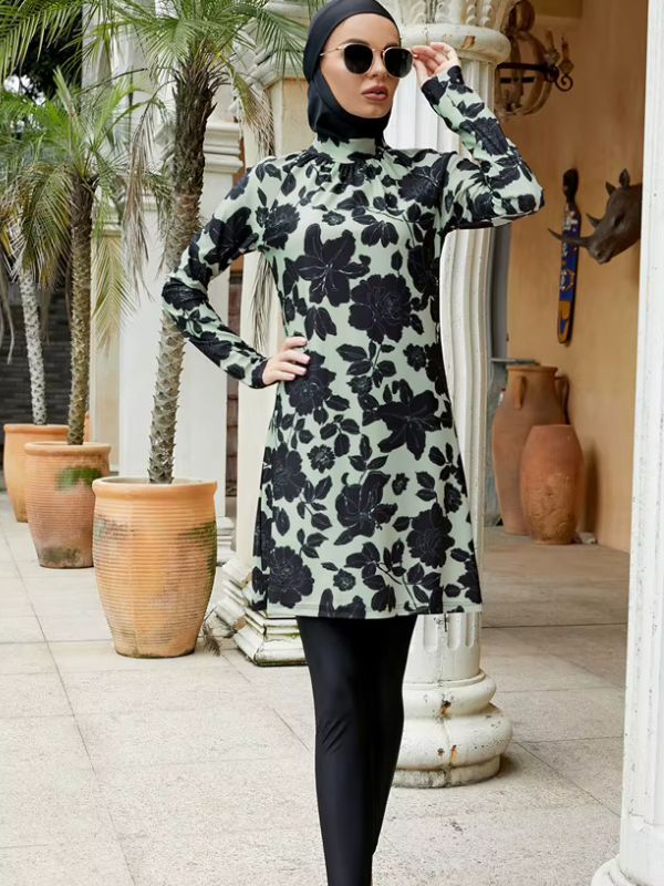 Burkini long vert sauge à grandes fleurs noires, vue de face avec lunettes de soleil et main sur la hanche, dans un décor de terrasse