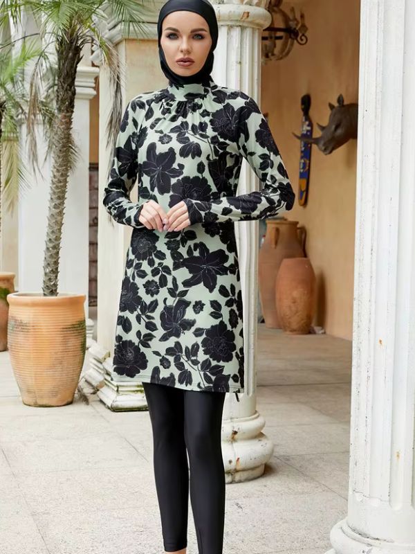 Burkini long vert sauge à grandes fleurs noires, vue de face avec lunettes de soleil et main sur la hanche, dans un décor de terrasse