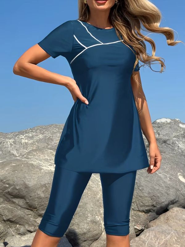 Burkini manche courte bleu, main sur la hanche, liséré blanc sur la poitrine, tunique évasée et legging bleu devant des rochers
