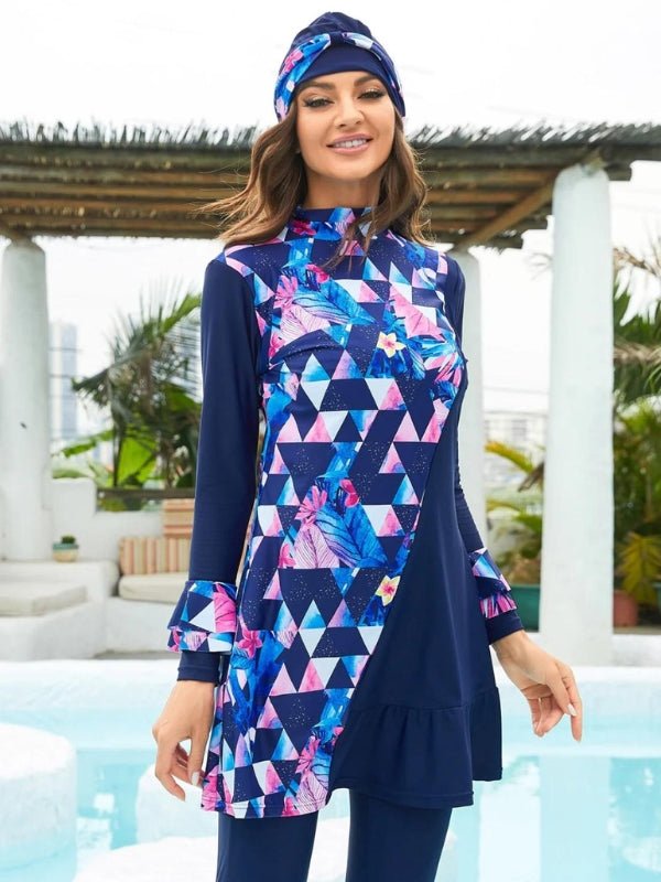 Une femme souriante porte un burkini bleu marine avec un motif géométrique audacieux de triangles roses et bleus, accentué de motifs floraux éclatants. La coupe élégante de la tenue, rehaussée par un volant subtil à l'ourlet, lui confère une allure à la fois moderne et féminine. Le turban assorti, décoré des mêmes motifs, complète harmonieusement l'ensemble, encadrant son visage avec style. Elle se tient près d'une piscine, ajoutant une touche de fraîcheur et de vacances