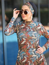 Une femme porte un burkini à motifs géométriques aux tons chauds et terreux, avec des touches de bleu, rouge, beige et noir. Elle a assorti son turban au même motif, créant ainsi une harmonie visuelle dans son look. Elle ajuste ses lunettes de soleil, mettant en valeur ses boucles d&#39;oreilles dorées. Son attitude confiante et élégante est soulignée par la main posée sur sa hanche