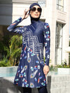 Une femme porte un burkini bleu marine avec un design distinctif composé de plumes de paon et de motifs floraux multicolores. Le motif comporte également des lignes ondulées blanches sur la partie supérieure de la tunique, créant un effet visuel attrayant. Elle porte un hijab assorti, également bleu marine, et des lunettes de soleil qui ajoutent une touche de sophistication