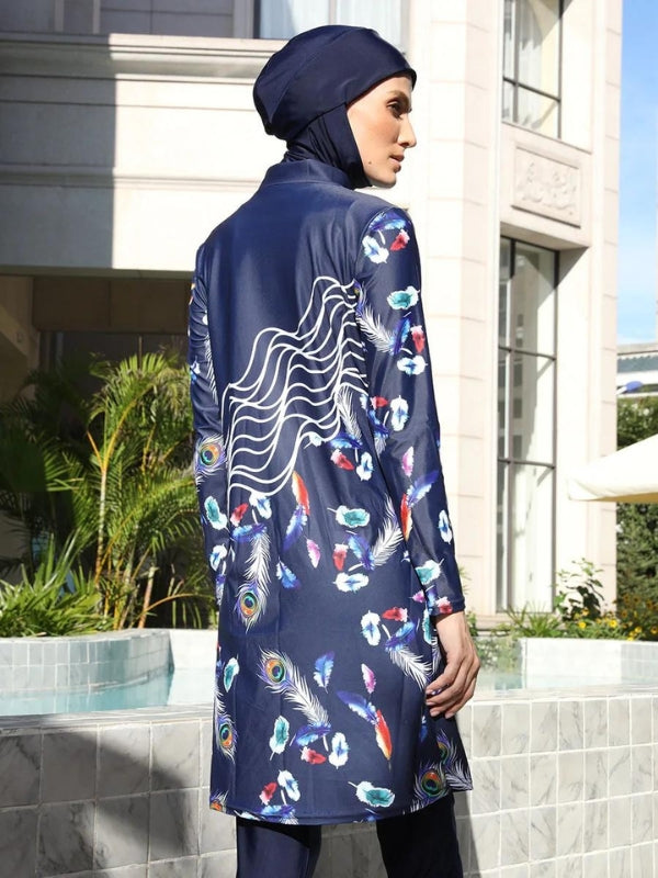 Une femme porte un burkini bleu marine avec un design distinctif composé de plumes de paon et de motifs floraux multicolores. Le motif comporte également des lignes ondulées blanches sur la partie supérieure de la tunique, créant un effet visuel attrayant. Elle porte un hijab assorti, également bleu marine, et des lunettes de soleil qui ajoutent une touche de sophistication