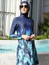 Une femme porte un burkini bleu marine orné de motifs abstraits dans des tons de bleu plus clair. Le design, qui s&#39;étend des manches jusqu&#39;à l&#39;ourlet de la tunique, crée un effet dynamique et moderne. Le hijab assorti et de grandes lunettes de soleil complètent cette tenue, offrant à la fois protection et style. Elle se tient avec assurance, le cadre élégant en arrière-plan mettant en valeur la sophistication de l&#39;ensemble