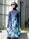 Une femme porte un burkini bleu marine avec des motifs abstraits en bleu clair sur la partie inférieure de la tunique, créant un effet artistique et moderne. Le hijab assorti et les grandes lunettes de soleil ajoutent une touche de sophistication à l&#39;ensemble. Elle se tient fièrement, avec une expression sereine, dans un cadre élégant qui met en valeur l&#39;élégance de sa tenue