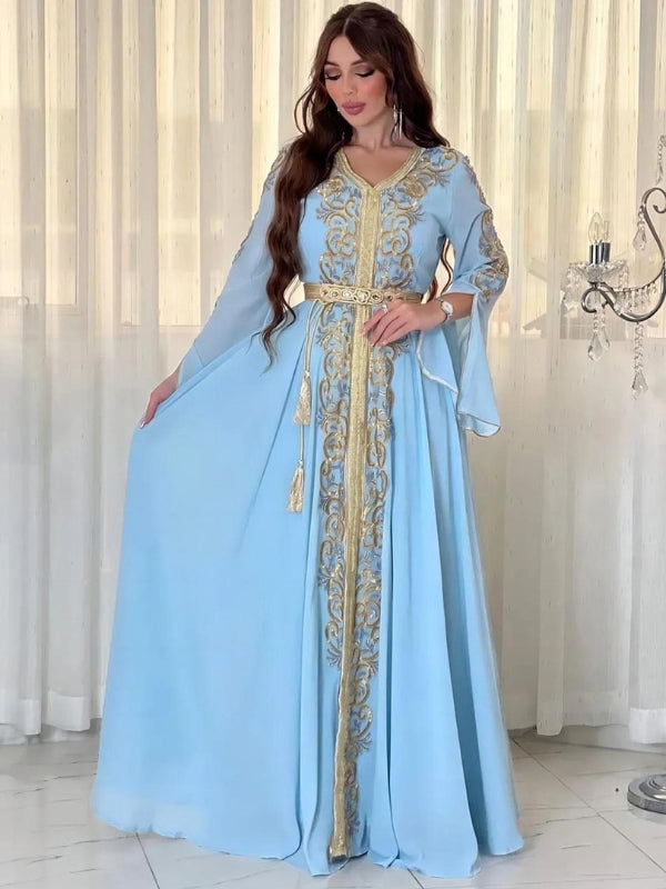 Une femme porte un caftan bleu clair somptueusement orné de motifs dorés élaborés qui descendent en ligne centrale de la robe, ainsi que sur les manches. La tenue est cintrée à la taille par une ceinture assortie, également décorée de détails dorés et agrémentée de pompons délicats. Le tissu fluide et léger tombe en plis élégants jusqu'au sol, offrant une allure majestueuse et gracieuse