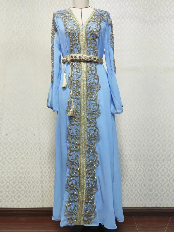 Une femme porte un caftan bleu clair somptueusement orné de motifs dorés élaborés qui descendent en ligne centrale de la robe, ainsi que sur les manches. La tenue est cintrée à la taille par une ceinture assortie, également décorée de détails dorés et agrémentée de pompons délicats. Le tissu fluide et léger tombe en plis élégants jusqu'au sol, offrant une allure majestueuse et gracieuse