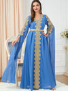 Femme vêtue d&#39;un caftan bleu roi richement brodé de motifs dorés le long de l&#39;encolure en V et descendant en ligne droite au centre de la robe. Les manches longues et amples ajoutent une touche de grâce au vêtement, qui est complété par une ceinture assortie soulignant la taille. Elle se tient debout dans une pièce élégante, posant avec confiance, et souriant légèrement. La scène est éclairée par une lumière douce, mettant en valeur les détails luxueux du caftan