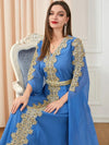 Femme assise, vêtue d&#39;un élégant caftan bleu roi avec des broderies dorées élaborées qui décorent le devant et les manches. Le caftan est agrémenté d&#39;une ceinture dorée qui met en valeur la taille. La femme porte des boucles d&#39;oreilles longues et scintillantes et un collier délicat. Ses cheveux bruns sont lisses et tombent sur ses épaules, tandis qu&#39;elle affiche un sourire subtil, créant une allure gracieuse et sophistiquée