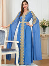 Une femme se tient debout, habillée d&#39;un caftan bleu roi somptueusement orné de broderies dorées élaborées, qui courent le long de l&#39;ouverture frontale, des manches et autour du décolleté. Le caftan est ajusté à la taille par une ceinture dorée, et ses manches longues et fluides ajoutent une touche majestueuse à l&#39;ensemble. Ses cheveux sont lisses, tombant sur ses épaules, et elle porte des boucles d&#39;oreilles pendantes. Elle sourit légèrement, posant avec élégance dans un intérieur chic