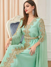 La femme est assise sur un fauteuil, portant un caftan vert menthe orné de broderies dorées. La ceinture assortie souligne sa taille, ajoutant une touche élégante à sa tenue. Ses longues boucles d&#39;oreilles et son collier complètent son look sophistiqué. Ses cheveux tombent en cascade sur ses épaules, et son maquillage est soigné, avec un rouge à lèvres qui ajoute une touche de couleur à son visage