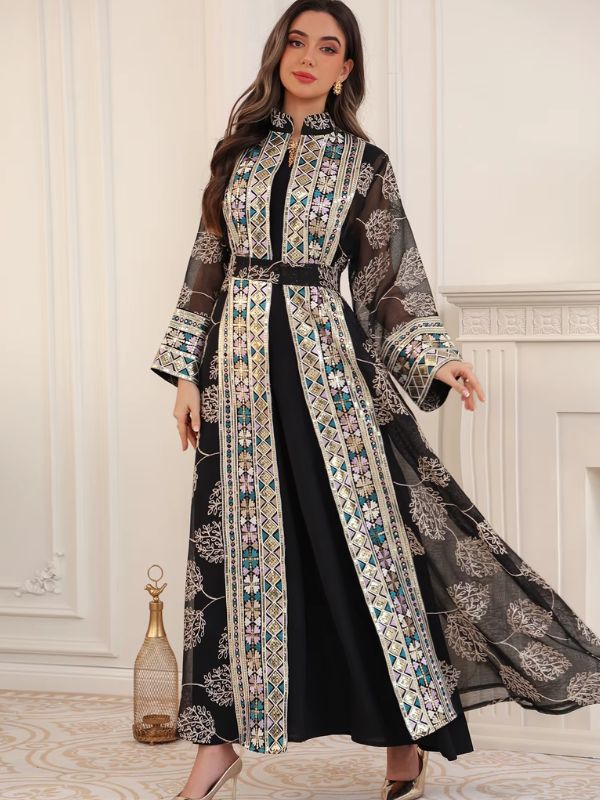 Femme portant un caftan noir fleuri à broderies géométriques, tenant un sac cage doré, devant une cheminée blanche.