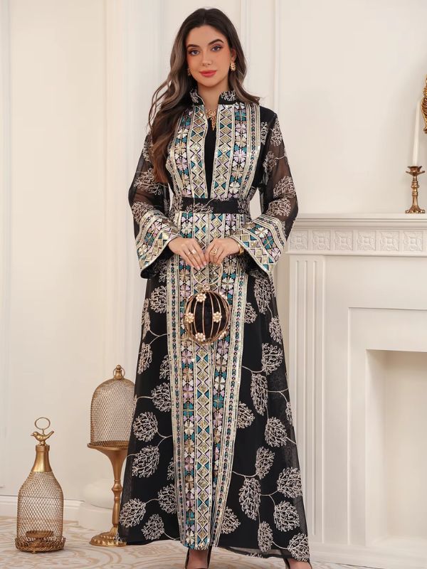 Femme portant un caftan noir en tulle fleuri, avec bandes brodées multicolores, près d'une cheminée.