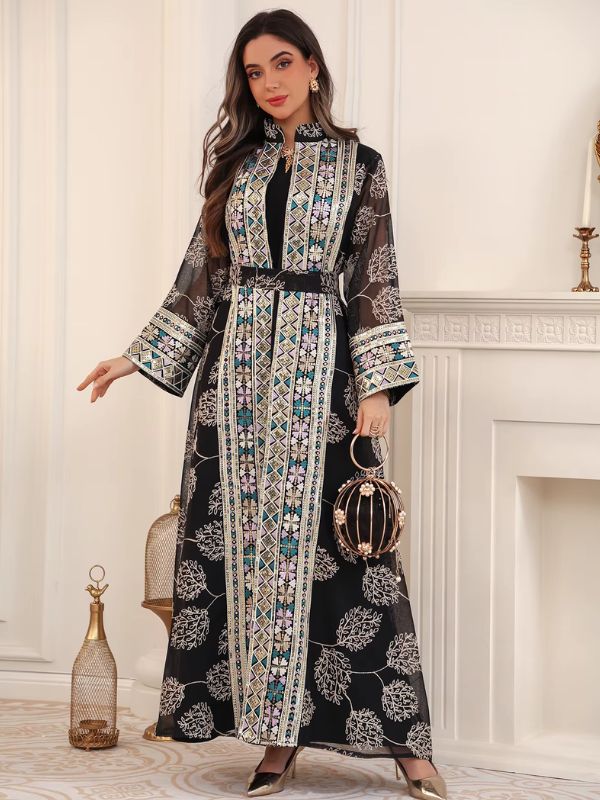 Femme portant un caftan noir fleuri à broderies géométriques, tenant un sac cage doré, devant une cheminée blanche.