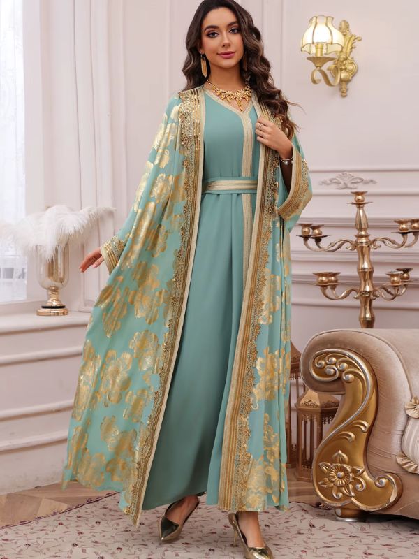 Femme en caftan turquoise floral doré, porté ouvert sur une robe assortie, près d'un chandelier doré