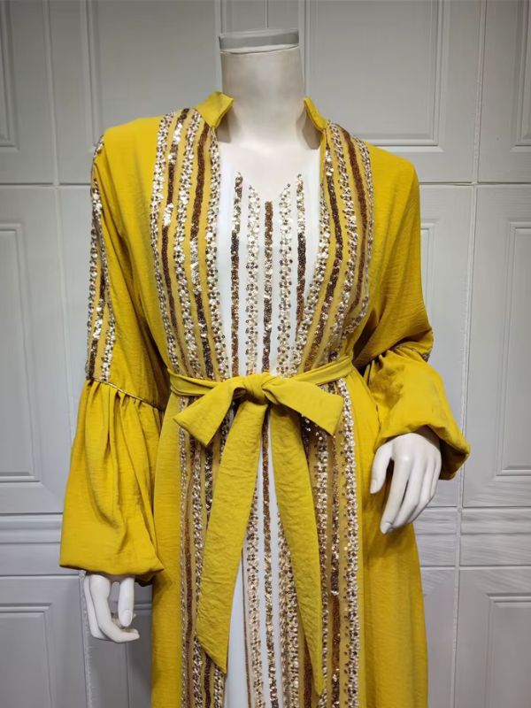 Femme portant un caftan jaune à manches ballon et sequins dorés sur une robe longue blanche, dans un décor élégant.