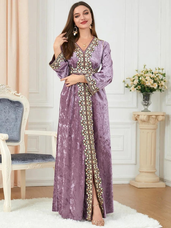 Une femme porte un caftan en velours mauve, orné de broderies dorées, noires et vertes le long du col en V, de la ligne centrale et des poignets. Elle a les cheveux bruns lâchés et porte des boucles d'oreilles dorées. Elle se tient dans un intérieur élégant avec un fauteuil blanc à coussin gris et une table ornée de fleurs. Ses talons dorés complètent son look sophistiqué