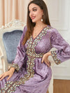 Une femme assise, portant un caftan en velours mauve orné de broderies colorées et complexes le long du décolleté, des manches et de la taille. Le caftan a un aspect luxueux et élégant, avec une texture riche en velours. La femme sourit doucement, avec des bijoux dorés, dans un cadre élégant avec une chaise ornée et un décor classique en arrière-plan