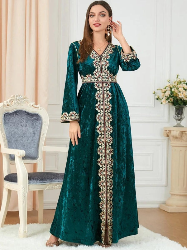 Une femme se tient debout, vêtue d'un magnifique caftan en velours vert émeraude, richement orné de broderies géométriques en rouge, or et beige qui décorent le col en V, descendent le long de la ligne centrale, et bordent les poignets des manches longues et évasées. Le caftan est cintré à la taille, soulignant élégamment la silhouette de la femme. La texture du velours apporte une profondeur luxueuse au vêtement, avec une légère brillance qui capte subtilement la lumière