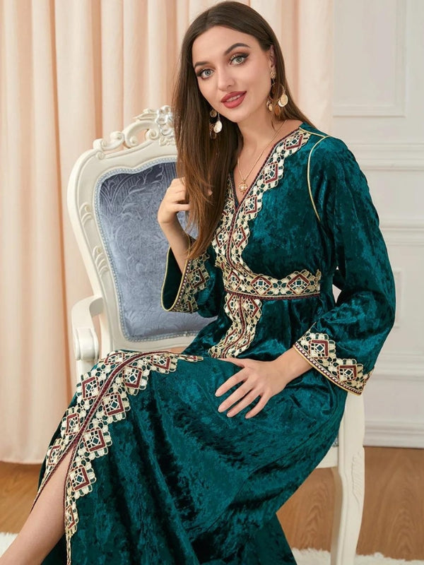 Une femme se tient debout, vêtue d'un magnifique caftan en velours vert émeraude, richement orné de broderies géométriques en rouge, or et beige qui décorent le col en V, descendent le long de la ligne centrale, et bordent les poignets des manches longues et évasées. Le caftan est cintré à la taille, soulignant élégamment la silhouette de la femme. La texture du velours apporte une profondeur luxueuse au vêtement, avec une légère brillance qui capte subtilement la lumière