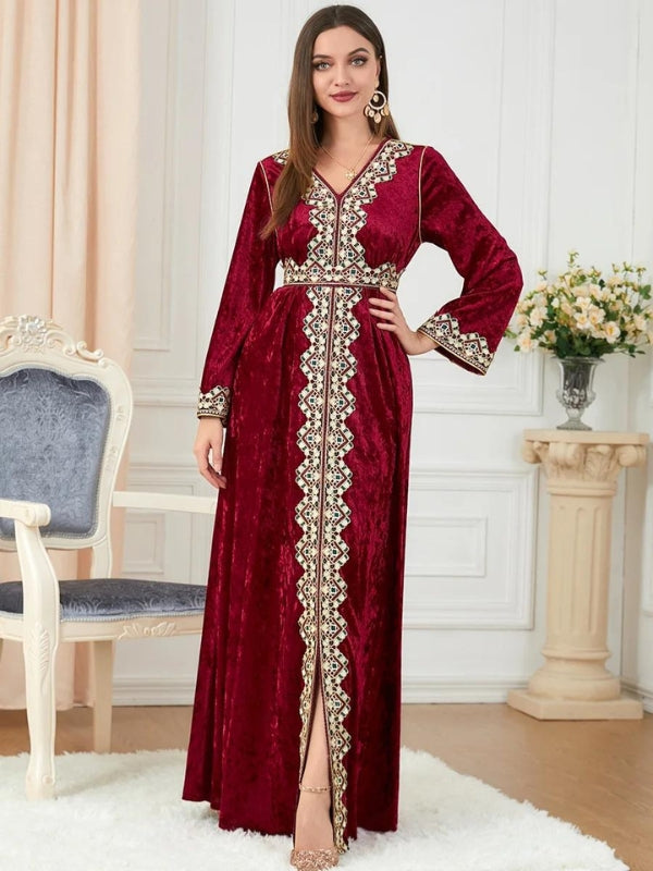 Une femme porte un caftan en velours rouge richement brodé, avec des détails dorés sur le devant, les manches, et autour de la taille. Le vêtement est long et élégant, fendu sur le devant, mettant en valeur ses chaussures à talons dorés. Elle se tient droite, la main posée sur sa hanche, avec une expression confiante. Ses cheveux longs tombent sur ses épaules, et elle complète son look avec de grandes boucles d'oreilles. L'ensemble crée une impression de sophistication et de glamour