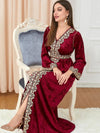 La femme est assise, portant un caftan en velours rouge avec des broderies dorées sur le devant et les manches. Sa posture met en avant la coupe élégante de la robe, tandis que ses chaussures dorées et ses bijoux assortis ajoutent une touche de glamour. Le contraste entre la couleur riche du caftan et les détails dorés crée un ensemble sophistiqué et raffiné