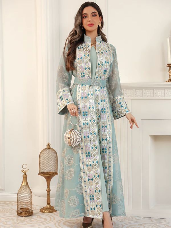 Femme portant un caftan turquoise brodé de motifs andalous, bras écartés, devant une cheminée blanche.