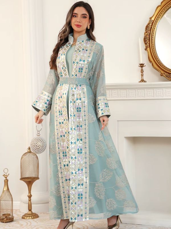 Femme portant un caftan turquoise en tissu floral brodé, avec bandes géométriques pastel, près d'une cheminée.