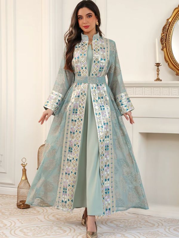 Femme portant un caftan turquoise brodé de motifs andalous, bras écartés, devant une cheminée blanche.