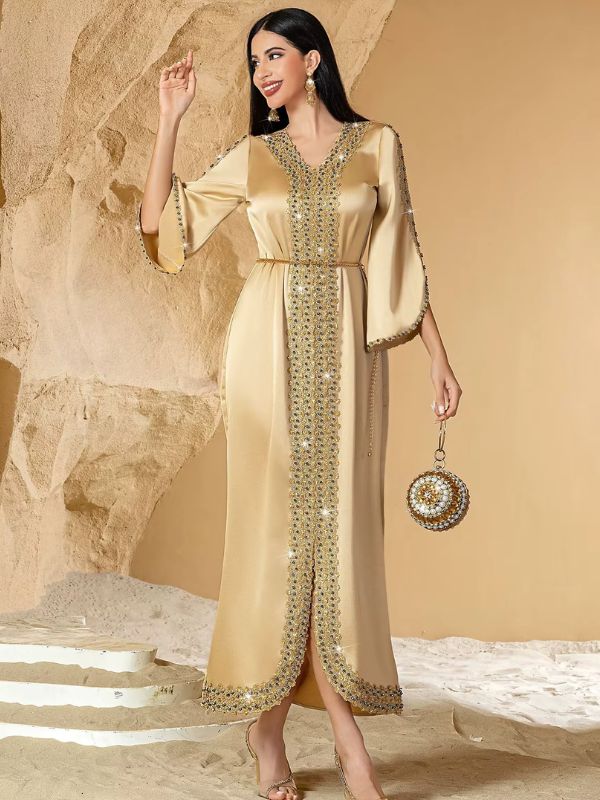 Caftan doré satiné à broderie de strass et pochette ronde assortie, posé devant des rochers de sable
