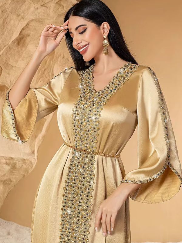 Caftan doré satiné à broderie de strass et pochette ronde assortie, posé devant des rochers de sable