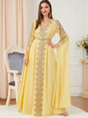 Une femme élégante porte un caftan jaune lumineux, richement décoré de broderies argentées qui ornent le centre et les manches de la robe. La tenue est ceinturée à la taille, accentuant sa silhouette gracieuse. Elle se tient debout avec confiance, une main sur la hanche, affichant un léger sourire. En arrière-plan, une chaise classique en bois blanc avec un tissu bleu et un vase de fleurs blanches ajoutent une touche de raffinement à la scène