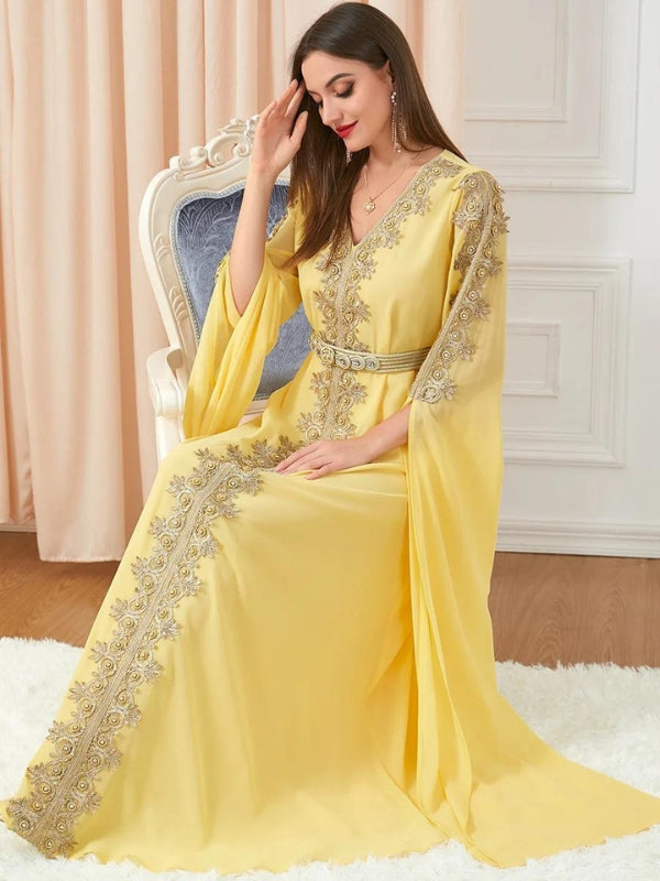 Une femme élégante porte un caftan jaune lumineux, richement décoré de broderies argentées qui ornent le centre et les manches de la robe. La tenue est ceinturée à la taille, accentuant sa silhouette gracieuse. Elle se tient debout avec confiance, une main sur la hanche, affichant un léger sourire. En arrière-plan, une chaise classique en bois blanc avec un tissu bleu et un vase de fleurs blanches ajoutent une touche de raffinement à la scène