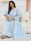  Une femme est assise, vêtue d&#39;un magnifique caftan bleu pastel. Le caftan, richement décoré, est cintré à la taille par une ceinture assortie qui met en valeur sa silhouette. Les manches longues et légèrement transparentes ajoutent une touche de légèreté à l&#39;ensemble. Elle porte des boucles d&#39;oreilles dorées qui ressortent joliment avec ses longs cheveux noirs ondulés