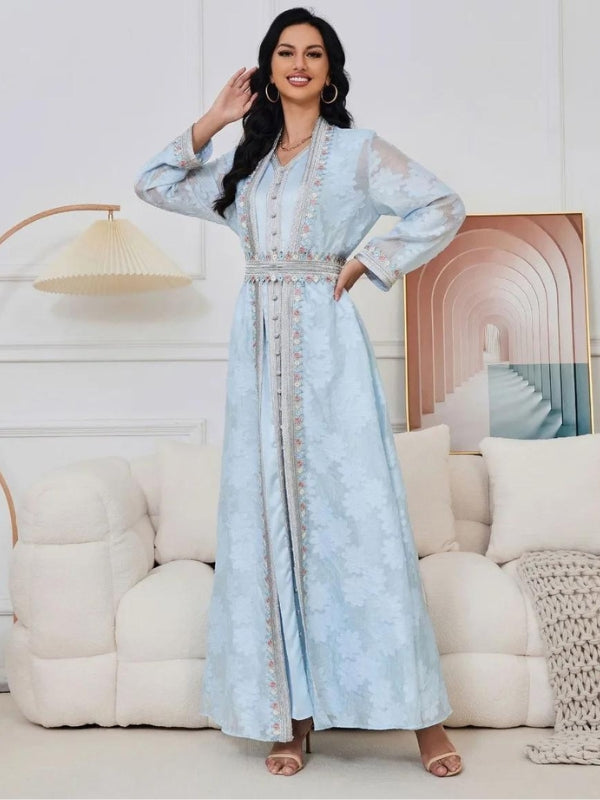 La femme sur cette image porte un caftan bleu pastel, orné de broderies délicates qui suivent la ligne centrale de la robe ainsi que les poignets. Le caftan est ajusté à la taille par une ceinture coordonnée, mettant en valeur sa silhouette. Le tissu semble léger et fluide, avec des motifs floraux subtils qui ajoutent une touche d&#39;élégance et de sophistication à l&#39;ensemble