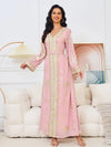 La femme porte un élégant caftan rose pastel, décoré de motifs dorés qui ajoutent une touche de sophistication à l&#39;ensemble. Le caftan est ajusté à la taille avec une ceinture qui accentue la silhouette tout en laissant tomber la jupe de manière fluide. Le col en V et les manches longues, également ornés de dorures, apportent une harmonie dans les détails