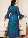 La femme sur cette image est vue de dos, portant un caftan bleu avec des motifs floraux rouges. Le caftan a des manches évasées et une ceinture qui se noue à l&#39;arrière, accentuant la taille. Les motifs floraux sont bien visibles, ajoutant une touche de vivacité à la tenue. Les bordures dorées et les détails délicats du caftan sont également visibles sur les manches