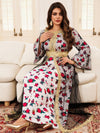 La femme est assise sur un canapé clair, portant un magnifique caftan avec un motif floral en rouge et vert sur fond blanc, recouvert d&#39;une sur-robe en tissu transparent gris à motifs floraux rouges, bordée de doré. La ceinture dorée à la taille met en valeur sa silhouette, et les manches évasées ajoutent une touche de sophistication. Ses cheveux longs et ondulés encadrent son visage, et elle complète son look avec de grandes boucles d&#39;oreilles dorées